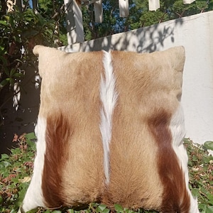May include: A square decorative pillow with a natural hide cover. The pillow features a central stripe of white fur, flanked by areas of tan and brown fur. The pillow is approximately 18 inches square, with a soft, textured appearance.