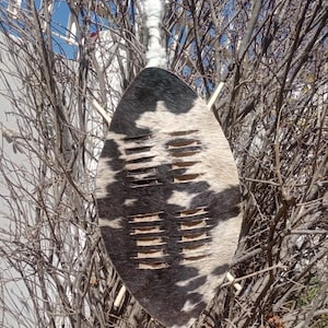 Puede incluir: Un escudo tradicional con un patrón de piel de vaca en blanco y negro. El escudo tiene forma de lágrima con ranuras horizontales y una parte superior blanca y esponjosa. Se muestra sobre un fondo de ramas de árboles desnudas y cielo azul.