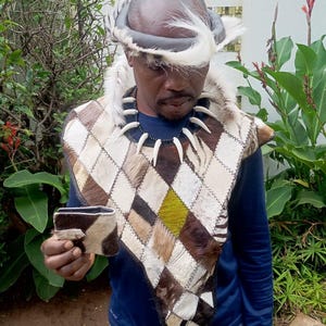 Puede incluir: Una persona con vestimenta africana tradicional, que incluye un chaleco de piel estampada, un collar de cuentas con dientes y un tocado de plumas. La persona sostiene una pequeña cartera. El chaleco presenta un patrón de diamantes con parches marrones, blancos y verdes.