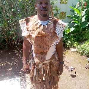 May include: A traditional African outfit featuring a brown and white spotted animal hide top and skirt. The outfit includes a matching headdress and a necklace with white teeth-like pendants. The person is wearing a white t-shirt underneath.