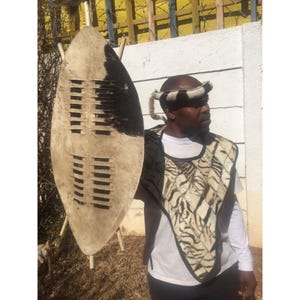 May include: A man wearing a traditional African garment made of animal hide, holding a shield made of cowhide with a wooden frame and leather straps. The shield has a pattern of holes cut into it.