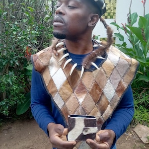 Puede incluir: Un atuendo tradicional con un chaleco de piel con estampado de diamantes en tonos marrones y beige, combinado con una cartera a juego. El conjunto incluye un collar con detalles blancos y un sombrero a rayas. La estética general es rústica y cultural.