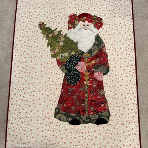 May include: A Christmas-themed quilt featuring a Santa Claus figure holding a Christmas tree. The Santa Claus is dressed in a red and green patterned coat and a red hat with a floral wreath. The background is a cream color with red floral accents.