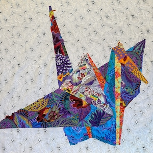 Może przedstawiać: Kolorowy żuraw origami wykonany z wzorzystej tkaniny. Żuraw jest złożony w tradycyjnym stylu i ma długą szyję i dziób. Tło to biała tkanina z powtarzającym się wzorem małych kółek.