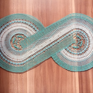 Può includere: Un delicato pizzo a tombolo fatto a mano in una combinazione di colori verde chiaro e bianco. Il pizzo ha la forma di un simbolo di infinito, con motivi intricati e una texture delicata.