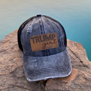 May include: A black and blue distressed denim trucker hat with a brown leather patch that says "TRUMP girl" on it.