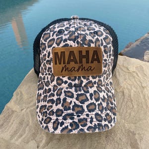 May include: A black and brown leopard print trucker hat with a brown leather patch that says "MAHA mama" in a brown font.