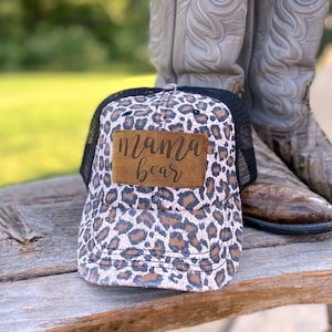 May include: A pink and black leopard print trucker hat with a brown leather patch that says "mama bear".