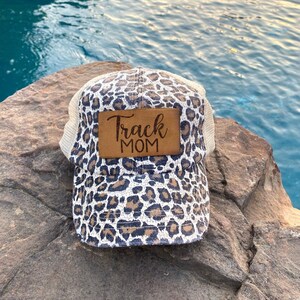 May include: A brown and white leopard print trucker hat with a brown leather patch that says "Track Mom" on it.