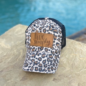May include: A black and brown leopard print trucker hat with a brown leather patch that says "live simply".