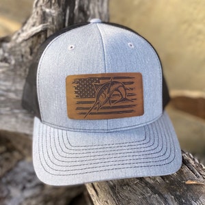 May include: A gray and black trucker hat with a brown leather patch featuring a marlin fish jumping over an American flag.