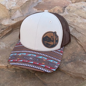 May include: A brown and white trucker hat with a brown leather patch featuring a cactus and mountain scene. The hat has a brown mesh back and a patterned brim with geometric designs in shades of brown, red, blue, and white.