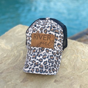 May include: A black and white leopard print trucker hat with a brown leather patch that says "River girl".