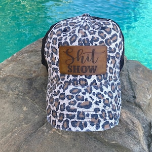 May include: A black and white leopard print trucker hat with a brown leather patch that says "Shit Show".