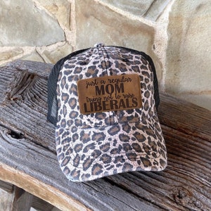 May include: A brown and black leopard print trucker hat with a brown leather patch that reads "just a regular MOM trying not to raise LIBERALS".