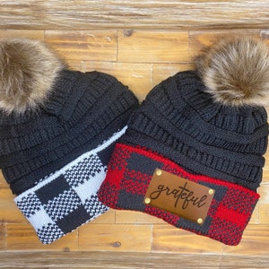 May include: Two black knit beanies with faux fur pom poms. One beanie has a black and white checkered cuff and the other has a red and black buffalo plaid cuff. The buffalo plaid beanie has a brown leather patch with the word "grateful" written on it.