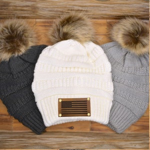 May include: Three knit beanies with faux fur pom-poms. One is white with a wooden American flag patch, another is dark gray, and the third is light gray. The beanies are displayed on a wooden surface.