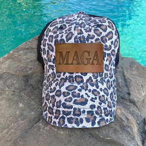May include: A black and white leopard print trucker hat with a brown leather patch that says "MAGA" on it.