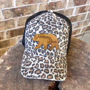 May include: A black and white leopard print trucker hat with a brown leather patch that says "Momma Bear" and a brown bear silhouette.