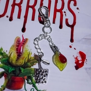 May include: A silver-coloured keychain with plant-themed charms. The charms include a small plant in a pot, a leaf-shaped charm with red details, and a chain. The background has the word "HORRORS" in red.