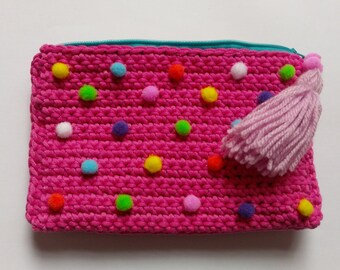 May include: A handmade pink crochet pouch with colorful pom-poms and a light pink tassel. The pouch has a turquoise zipper and is a rectangular shape. This item is perfect for storing small items.