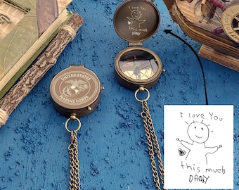 May include: A brass compass with a chain, featuring a United States Marine Corps emblem on the front and a personalised message on the back. The message reads "I love you this much DADDY" with a stick figure drawing.