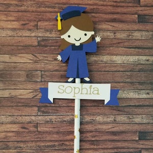 May include: A blue and white graduation cake topper with a girl wearing a blue cap and gown. The topper has a banner that says "Sophia".