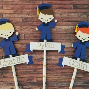May include: Three graduation cap toppers with blue gowns and yellow tassels. Each topper has a name on a banner: Charlie, Dylan, and Colin.