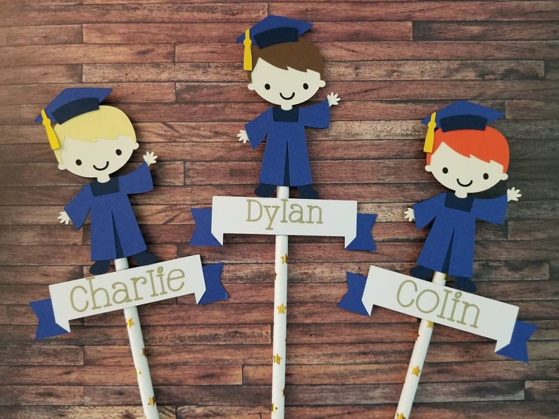 May include: Three graduation cap toppers with blue gowns and yellow tassels. Each topper has a name on a banner: Charlie, Dylan, and Colin.