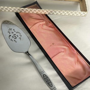 May include: A silver pie server with a decorative handle and a slotted blade, presented in a black box with a pink satin lining. The box has a clear lid and a patterned beige and white trim.