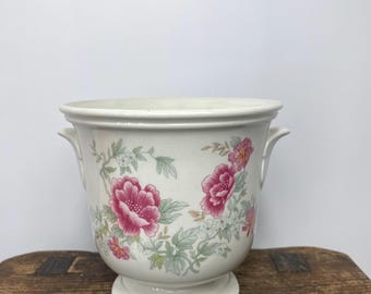 May include: Cream-colored ceramic planter with a pedestal base and two small handles. The planter is decorated with pink floral patterns and green foliage. A decorative container for plants.