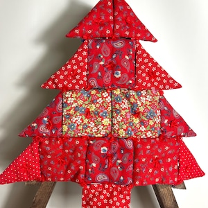 May include: A red quilted Christmas tree decoration with paisley and floral patterns. The tree is made from layered, triangular sections and adorned with small red bows. It is displayed on a wooden stand, adding a festive touch to any space.