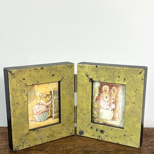 Peut inclure: Un cadre photo double articulé avec une finition vert olive vieilli et des bords brun foncé. Le cadre gauche contient une illustration d'une souris en veste rose ouvrant une porte, et le cadre droit montre une souris en veste rose debout.
