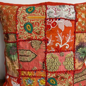 May include: A square throw cushion featuring a patchwork design of orange fabric panels. Each panel is decorated with intricate embroidery in gold, green, white, and red thread, incorporating floral motifs, geometric patterns, and metallic sequins.