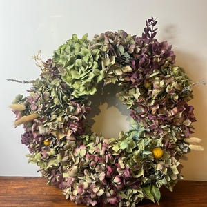 May include: A circular wreath composed of dried hydrangea flowers in shades of muted green and deep purple. The arrangement includes small yellow billy ball flowers, wispy beige grass plumes, and thin sprigs of dark purple foliage throughout.