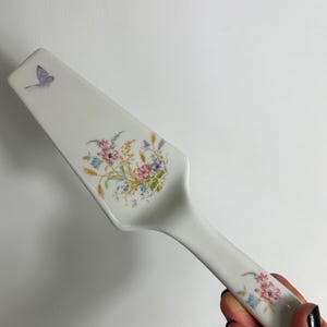 May include: White ceramic cake server with a floral design featuring pink, blue, and yellow flowers. A small purple butterfly is on the top corner. The handle has a curved shape for easy grip.