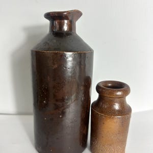 May include: Two cylindrical stoneware vessels with a dark brown glazed finish. The taller bottle features a narrow neck and a flared rim, while the shorter, wider jar has a thick, rounded rim and a lighter, textured base with visible speckling.
