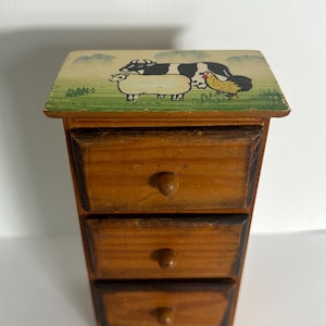 May include: A small wooden chest of drawers with three stacked drawers featuring round knobs. The top surface is painted with a green field scene depicting a black and white cow, a white sheep, and a brown chicken under a light blue sky.