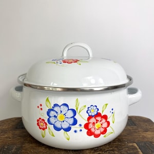 May include: A white enamel pot with a matching lid, featuring a floral pattern of blue and red flowers with green leaves. The pot has two side handles and a silver-toned metal rim around the top edge, with a rounded handle on the lid.