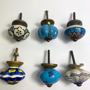 May include: Six ceramic cabinet knobs with brass fittings. The knobs display diverse designs, including floral patterns and solid colours like blue and white. Each knob has a unique shape and design, suitable for furniture.