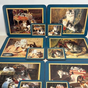 May include: A collection of eight placemats and coasters. Each item displays a detailed painting of cats in domestic scenes. The placemats and coasters have a dark teal border with a gold trim. The artwork shows cats on furniture and with books.