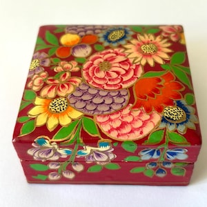 May include: A rectangular box with a deep red base color, featuring a hand-painted floral pattern across the lid and sides. The design includes various flowers in shades of pink, orange, yellow, blue, and purple, accented with green leaves.