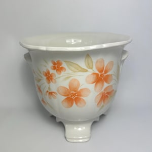 May include: A white ceramic planter with a floral design. The planter features orange flowers and green leaves painted on the exterior. It has a flared rim, two small handles, and a pedestal base. The overall shape is rounded.