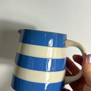 May include: A ceramic pitcher with a blue and cream striped pattern. The pitcher has a curved handle and a spout for pouring. The design is reminiscent of nautical themes. The pitcher is a decorative item.