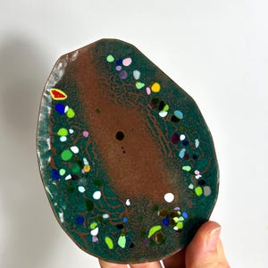 May include: An irregularly shaped, decorative dish with a dark brown center and a teal green border. The dish is speckled with colorful dots of white, yellow, green, blue, and purple. The dish is held by a hand.