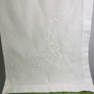 May include: White linen textile featuring embroidered floral and scroll detailing. The embroidery is in white thread, creating a subtle pattern. The fabric is folded over a patterned surface, with a stitched edge.