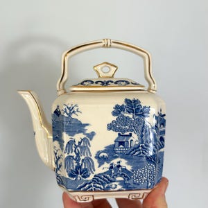 May include: Cream ceramic teapot with a blue and white landscape design. The teapot has a square shape, a curved spout, and a gold handle and trim. The lid features a small finial. The design depicts trees, buildings, and bridges, reminiscent of a classic Willow pattern.