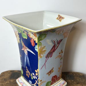 May include: A square porcelain vase featuring alternating panels of deep blue and light grey. The surface is decorated with colourful hummingbirds, butterflies, and floral motifs. The base is accented with a pink and white scalloped border design.