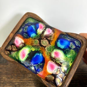 May include: A rectangular wooden trinket box with a colorful, abstract design. The box features a vibrant mosaic of colors, including blue, pink, green, and orange, set within a dark wooden frame. The design appears to be made of glass or resin.
