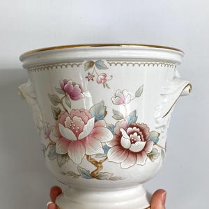 May include: White ceramic planter with a gold rim and handles. The planter is decorated with pink and brown floral designs and blue leaves. The planter has a pedestal base.